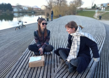 Nina and Patricija are sitting on the pier. In front of them is an unopened box with first protoypes of the cards.